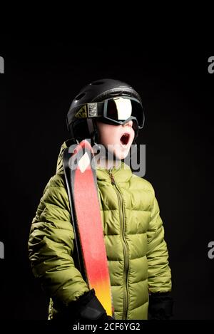 J'ai surpris un petit enfant en portant un casque et des lunettes de ski chaudes veste et gants et ski sur les épaules sur le noir arrière-plan en studio Banque D'Images