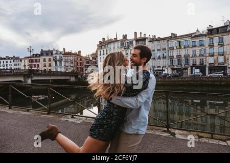 Vue latérale d'un jeune couple affectueux et heureux qui s'embrasse et rit en vous tenant sur un remblai de pierre près de la rivière avec de vieux bâtiments En arrière-plan dans la ville de Bayonne en France Banque D'Images