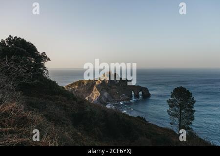 Chemin de la pierre de pavage menant le long du pont de pierre et de l'arête de rocky colline à la maison de campagne solitaire sur l'île Gaztelugatxe entouré par l'eau de mer tranquille avec des vagues de mousse blanche Banque D'Images