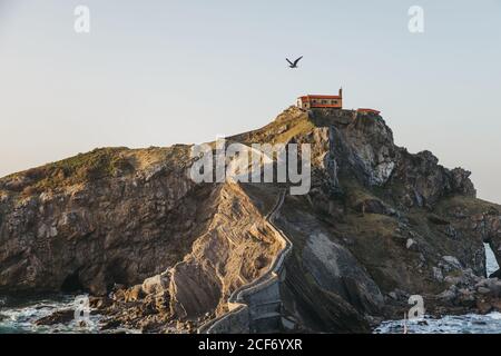 Chemin de la pierre de pavage menant le long du pont de pierre et de l'arête de rocky colline à la maison de campagne solitaire sur l'île Gaztelugatxe entouré par l'eau de mer tranquille avec des vagues de mousse blanche Banque D'Images