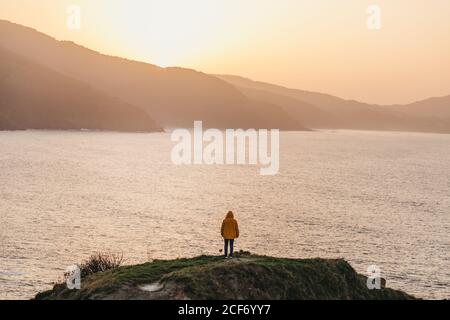 Vue arrière d'un homme méconnaissable dans une veste jaune vif et jean se tenant sur une colline rocheuse et appréciant le paysage pittoresque de côte de mer au coucher du soleil en Espagne Banque D'Images