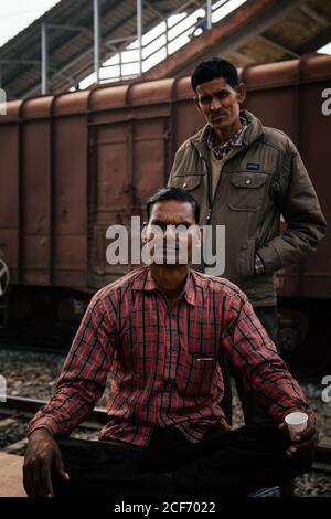 Allahabad City, Inde - FÉVRIER 2018 : un Indien sérieux adulte en chemise à carreaux avec une tasse de boisson à la main assis avec des jambes croisées sur les rails tandis qu'un collègue en veste se tient derrière et les deux regardant la caméra avec chariot de transport en arrière-plan Banque D'Images