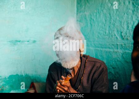 Varanasi, Inde - février, 2018: Pauvre homme âgé hindou en bonnet tricoté beige assis dans le coin de la maison avec des murs bleus et fumage cigare Banque D'Images