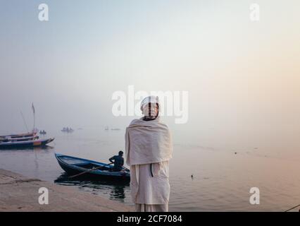 Varanasi, Inde - FÉVRIER, 2018: Homme indien âgé portant des vêtements blancs traditionnels debout sur la rive de la rivière avec des bateaux de pêche sur l'eau tôt le matin brumeux Banque D'Images