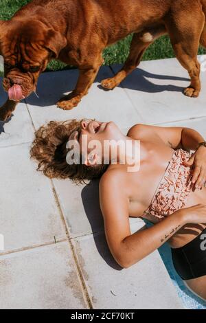 Par-dessus une femme bronzée joyeuse en maillot de bain riant en étant couché sur le sol et en jouant avec un grand chien brun par beau temps Banque D'Images