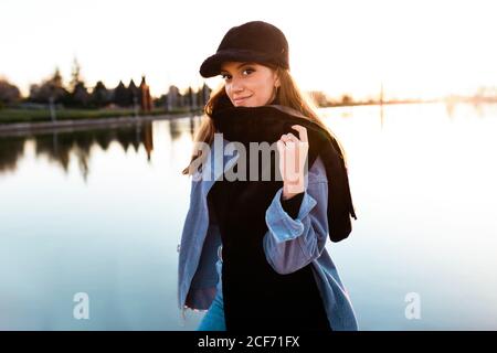 Charmante jeune femme en casquette noire et foulard souriant appareil photo avec lac tranquille sur fond flou au coucher du soleil Banque D'Images