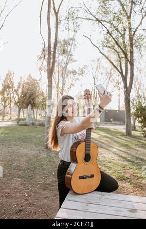 Jolie jeune femme avec guitare acoustique souriant gaiement et posant pour selfie tout en se tenant près de la table le jour ensoleillé dans le parc Banque D'Images