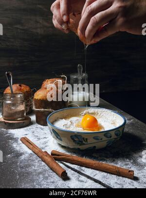 Crop personne faisant le panettone traditionnel et casser l'oeuf en céramique bol placé sur une table en bois avec de la cannelle et de la vanille et gâteaux cuits prêts à cuire en arrière-plan Banque D'Images