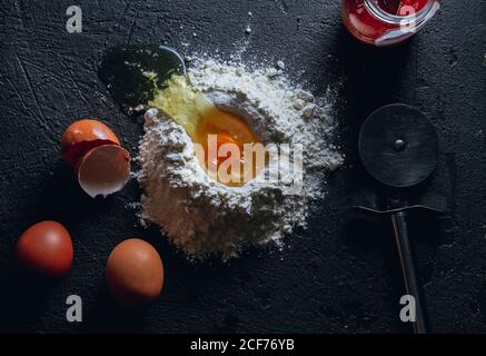 Vue de dessus de l'œuf cassé dans la farine sur le noir texturé surface avec couteau rond pour couper la pâte et le pot en verre Banque D'Images