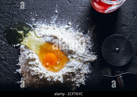 Vue de dessus de l'œuf cassé dans la farine sur le noir texturé surface avec couteau rond pour couper la pâte et le pot en verre Banque D'Images