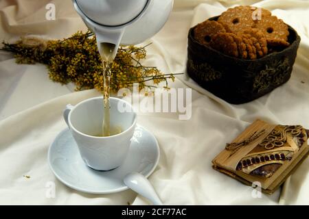 Théière blanche versant du thé vert dans une tasse placée sur le lit avec petits gâteaux pour un délicieux petit déjeuner Banque D'Images