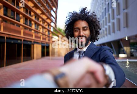 Point de vue prise de vue d'un homme d'affaires ethnique souriant vêtements formels secouant la main floue de l'associé d'affaires et de regarder à l'appareil photo Banque D'Images