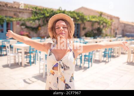 Attirant une femme adulte en chapeau de paille portant une robe légère avec les yeux fermés et les bras ouverts tout en étant debout seul sur le café terrasse avec mobilier bleu et blanc en plein soleil Banque D'Images