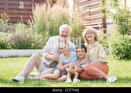 Portrait de groupe de grand-mère, de grand-père et de deux petits-enfants, assis ensemble sur la pelouse dans l'arrière-cour, en regardant l'appareil photo sourire Banque D'Images