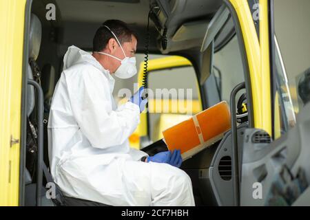 Vue latérale du pompier portant un uniforme de protection et des gants médicaux assis dans un feu moteur regardant l'ordinateur portable et parlant dessus talkie-walkie Banque D'Images