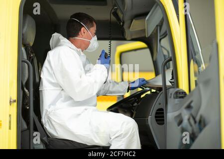 Vue latérale du pompier portant un uniforme de protection et des gants médicaux assis dans un feu moteur regardant l'ordinateur portable et parlant dessus talkie-walkie Banque D'Images