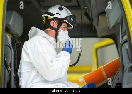 Vue latérale du pompier portant un uniforme de protection et un casque rigide avec des gants médicaux assis dans un véhicule d'incendie parlant sur l'émetteur-récepteur portatif talkie Banque D'Images