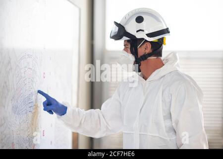 Vue latérale du pompier portant un costume de protection et un respirateur debout à la caserne de pompiers et en pointant sur le plan papier joint à mur Banque D'Images