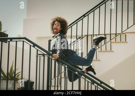 Vue latérale d'une femme afro-américaine excitée regardant la caméra et crier tout en se penchant sur les rampes et en sautant sur le bâtiment escaliers Banque D'Images