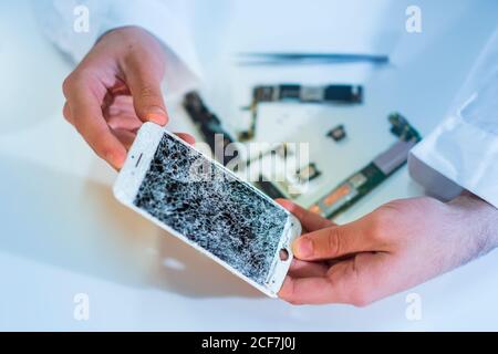 Mains d'un technicien méconnaissable qui présente un écran fissuré d'un mobile moderne téléphone sur table blanche dans l'atelier Banque D'Images