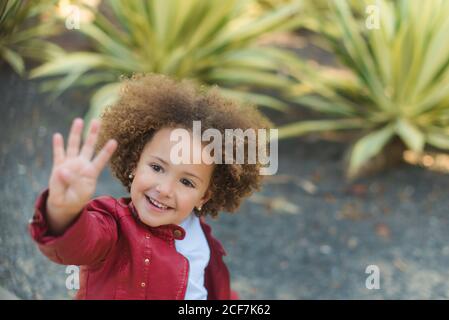 Grand angle de gaie petite fille aux cheveux bouclés en rouge veste montrant quatre doigts et souriant en se tenant contre le vert plantes tropicales dans le parc Banque D'Images