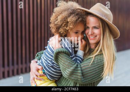 Vue latérale d'une jeune femme gaie dans un chandail tendance et chapeau enveloppant petite fille mignonne en se tenant dans la rue à proximité clôture moderne Banque D'Images