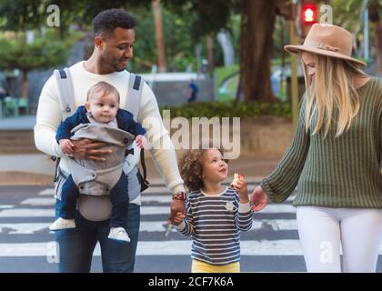 Heureux jeunes parents multiraciaux avec petite fille mangeant de la pomme et petit garçon en croisant la route sur le zèbre tout en marchant ville en été Banque D'Images