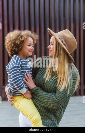 Vue latérale d'une jeune femme gaie dans un chandail tendance et chapeau enveloppant petite fille mignonne en se tenant dans la rue à proximité clôture moderne Banque D'Images