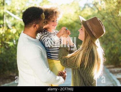 Joyeux jeune famille multiraciale avec un petit enfant qui profite du temps ensemble tout en se reposant dans un parc verdoyant par une belle journée d'été Banque D'Images