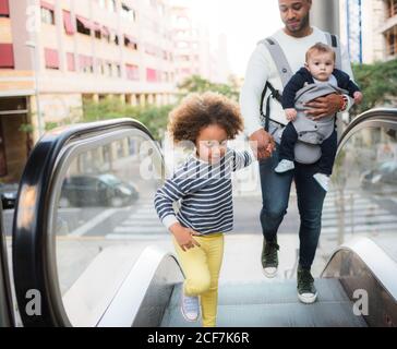 Par-dessus le jeune homme ethnique positif transportant un bébé fils et en tenant la main de la petite fille joyeuse tout en restant debout escalier roulant en ville Banque D'Images