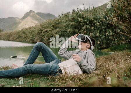 Jeune adolescent avec lunettes de réalité virtuelle posé sur l'herbe à l'extérieur près d'un lac avec un livre Banque D'Images