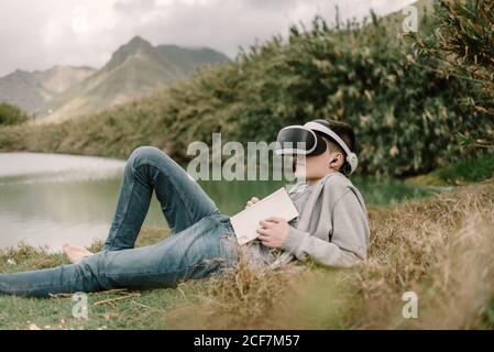 Jeune adolescent avec lunettes de réalité virtuelle posé sur l'herbe à l'extérieur près d'un lac avec un livre Banque D'Images