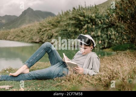 Jeune adolescent avec lunettes de réalité virtuelle posé sur l'herbe à l'extérieur près d'un lac avec un livre Banque D'Images