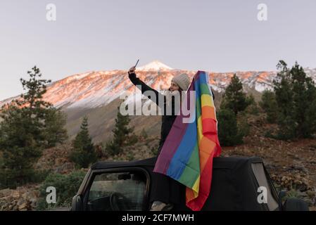 Femme active dans des vêtements chauds assise sur un SUV noir et prenant selfie sur téléphone mobile avec drapeau LGBT coloré en montagne à Tenefire, île des Canaries, Espagne Banque D'Images
