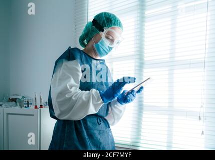 Vue latérale de la femme médecin spécialiste en robe de protection avec masque et lunettes de protection pour naviguer sur votre téléphone mobile et regarder fenêtre obturée pendant le travail à l'hôpital pendant l'éclosion du coronavirus Banque D'Images