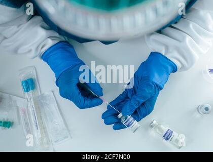 Du dessus de la récolte scientifique anonyme ou médecin en protection gants remplissant la seringue du flacon avec le vaccin coronavirus pendant le travail au tableau avec tubes à essai Banque D'Images