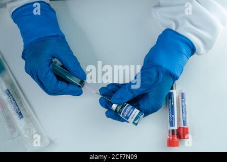 Du dessus de la récolte scientifique anonyme ou médecin en protection gants remplissant la seringue du flacon avec le vaccin coronavirus pendant le travail au tableau avec tubes à essai Banque D'Images