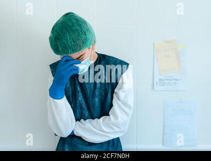 Épuisé jeune travailleur médical en costume de protection et masque avec gants se tenant près du mur dans le couloir de l'hôpital tout en travaillant dur au cours d'une pandémie de coronavirus Banque D'Images
