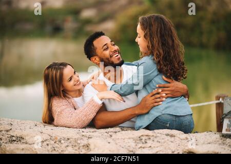Heureux décontracté parents multiraciaux couché sur terre de sable ensemble et regarder avec admiration le contenu de la fille assis à côté et embrassant père Banque D'Images