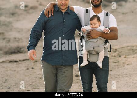 Petit noir décontracté homme tenant sur l'épaule anonyme homme petit ami et tenir le bébé calme dans un chariot gris tout en se promenant sur la nature en journée Banque D'Images