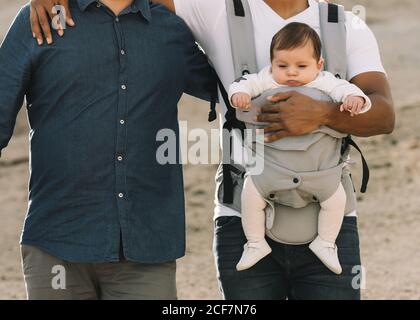 Petit noir décontracté homme tenant sur l'épaule anonyme homme petit ami et tenir le bébé calme dans un chariot gris tout en se promenant sur la nature en journée Banque D'Images