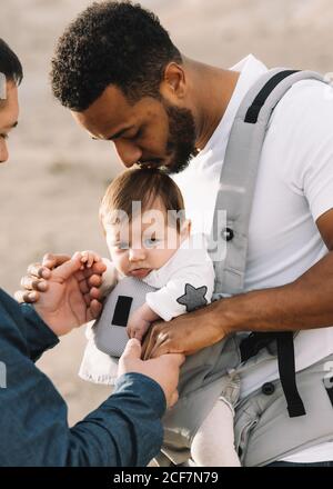 Vue latérale d'un homme décontracté noir tenant la main de l'anonyme petit ami masculin et garde un bébé calme dans un porteur gris tout en se promenant dans la nature pendant la journée Banque D'Images