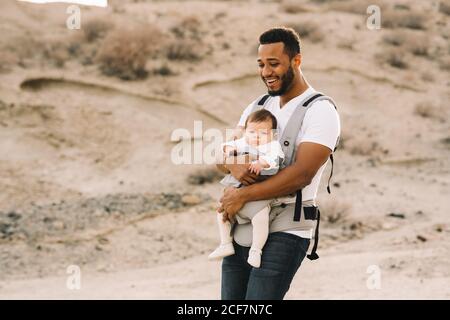 Joyeux homme américain africain barbu en t-shirt blanc et jeans tenant le nouveau-né dans le porte-bébé tout en se promenant sur la nature Banque D'Images