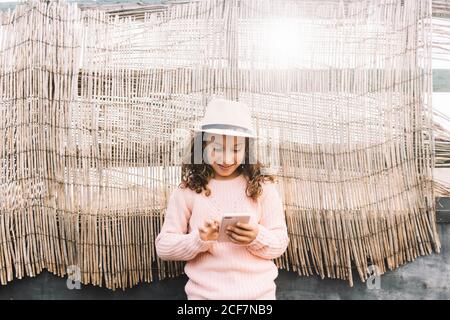 Fille concentrée en paille chapeau rose pull et jeans se tenir sur un fond de bois et naviguer sur un téléphone mobile Banque D'Images