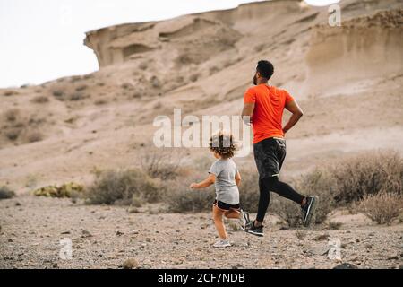 Père actif barbu afro-américain en t-shirt rouge courant avec un enfant gai sur un paysage désertique en contre-jour Banque D'Images
