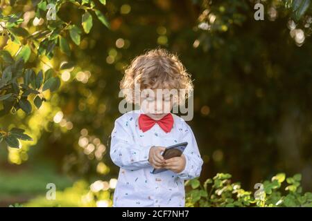 Petit garçon aux cheveux bouclés dans une chemise et un noeud papillon rouge utilisation d'un téléphone portable avec des plantes vertes sur un arrière-plan flou Banque D'Images