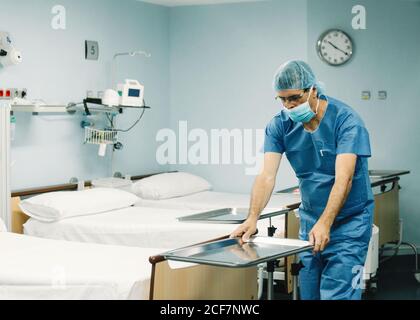 Medic en bleu uniforme et plateau de mise en place du masque de protection chariot dans la chambre d'hôpital à côté de lits vides Banque D'Images