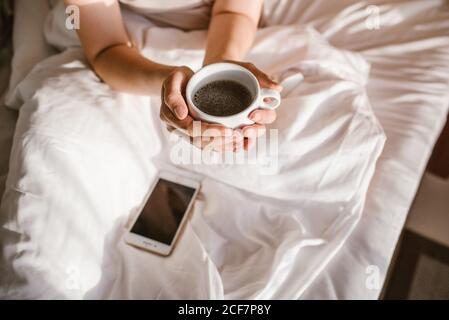 De dessus, crop une femme adulte méconnaissable qui interagit avec un smartphone pendant asseyez-vous dans le lit et tenez-vous tout en prenant une tasse de café ronde le matin ensoleillé Banque D'Images