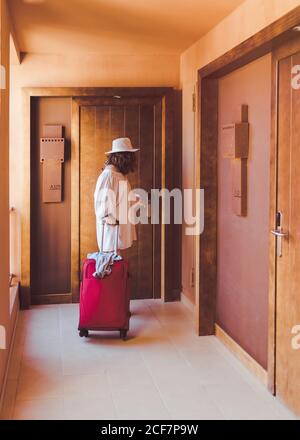 Femme adulte et vêtements blancs décontractés avec valise rouge fermant la porte en bois dans le couloir de l'hôtel Banque D'Images