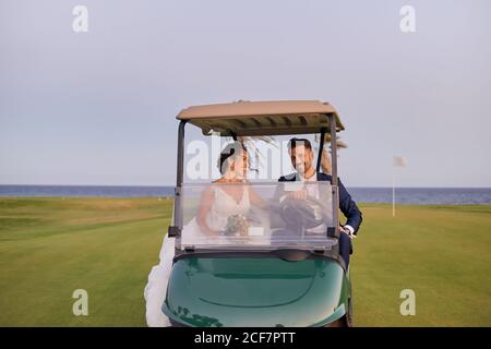 Mariée souriante avec bouquet de fleurs et beau marié combinaison élégante assise dans une voiturette de golf et traversant le green pelouse Banque D'Images